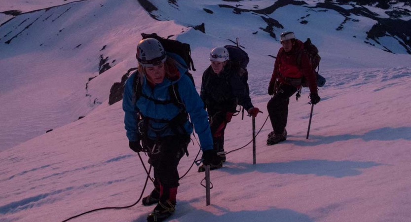 Three people wearing mountaineering gear are attached to each other by a rope. The snow appears pink and purple, presumably from the sunset or sunrise. 